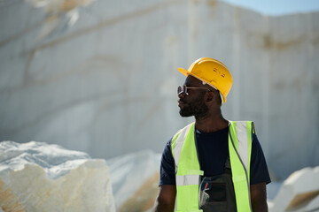 Aufkleber - Young black man in workwear, eyeglasses and protective helmet moving down territory of quarry and looking aside against marble rocks