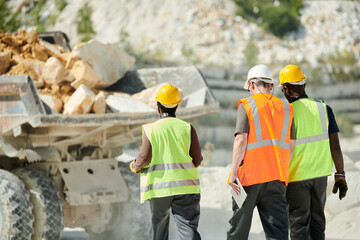 Aufkleber - Rear view of intercultural engineers in uniform and hardhats walking towards dump truck with heap of marble rocks in its body