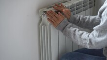 Closeup Of Woman In Sweater Warming Her Hands On The Heater At Home During Cold Winter Days. Female Getting Warm Up Her Arms Over Radiator. Concept Of Heating Season, Cold Weather. 