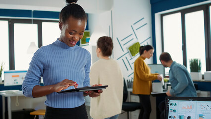 Wall Mural - Portrait of african american woman standing in busy office picking up tablet with business erp software and smiling looking at touchscreen. Employee holding digital device and interacting with screen.