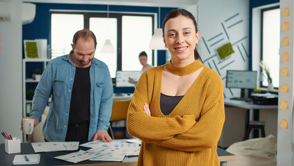 Wall Mural - Portrait of confident startup employee entering frame posing with arms crossed and smiling in busy modern office. Business woman acting casual and relaxed in financial department of small company.