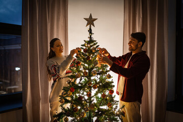 Wall Mural - winter holidays and people concept - happy couple decorating christmas tree at home