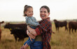 © Nina L/peopleimages.com - Family, mother and baby on a farm with cows in the background eating grass, sustainability and agriculture. Happy organic dairy farmer mom with her girl and cattle herd outside in sustainable nature