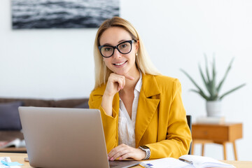 Portrait of successful young woman working with laptop. Businesswoman with glasses at work sitting at workplace in modern office looking at the camera and smiles friendly