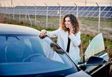Girl Talking On Phone While Standing Near Open Car And Looking To Left. On Backgdrop Solar Power Plant Producing Sustainable Electricity And Renewable Energy. Economic And Ecology Energy Indystry.