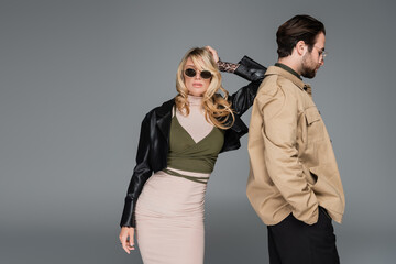 stylish blonde woman in sunglasses and leather jacket leaning on back of bearded man isolated on grey