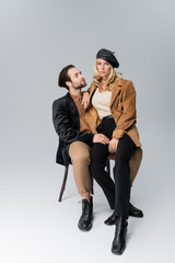 Wall Mural - full length of blonde woman in stylish beret sitting on knees of bearded man in sunglasses on grey