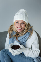 Wall Mural - happy blonde woman in winter outfit sitting in comfortable armchair and holding cup of coffee isolated on grey