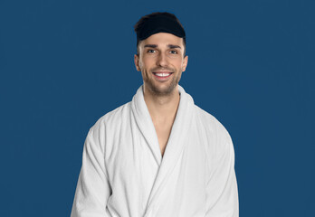 Happy young man in bathrobe and eye sleeping mask on blue background