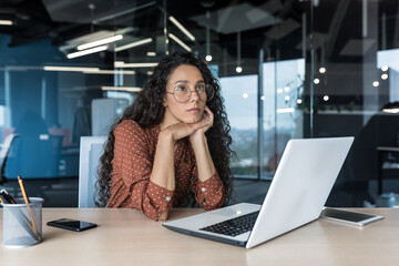 Wall Mural - Young beautiful hispanic woman with curly hair and glasses, working in modern office using laptop, female worker thinking and sad, business woman upset