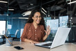 © Liubomir - Successful and beautiful hispanic woman working inside modern office building, businesswoman using laptop for video call smiling and waving, greeting gesture, online conference with colleagues
