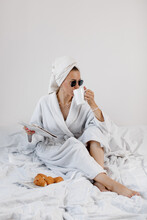 Happy Woman In A White Bathrobe And With A Towel On Her Head Drinks Coffee And Eats A Croissant In Bed In The Morning. Lifestyle Concept. 
