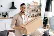 Joyful modern indian or arabian guy, stand at home in the living room, holding a large cardboard box, received parcel from the online store, preparing to unpack, looks at camera, smile, happy emotions