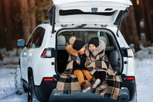 Two Happy Little Children Are Having Fun On A Winter Walk. Children Sit In Trunk Of Family Car. Children Warm Themselves Wrapped In Warm Blanket.