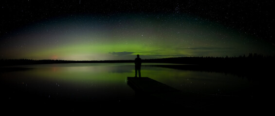 Wall Mural - person standing on a dock looking at a night sky filled with stars and aurora reflecting in calm wat