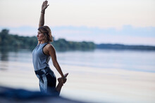 A Woman Is Practicing Yoga And Balancing On One Foot On The Dock Near The River In Nature.