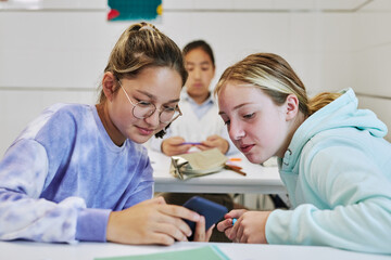 Wall Mural - Portrait of two teenage girls using smartphone together sharing social media or playing games in school classroom