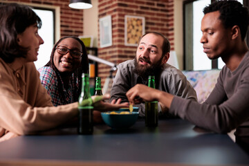 Wall Mural - Positive group of multiethnic friends sitting at table while discussing and having fun together. Multiracial people at home in living room enjoying snacks and beverages while talking.