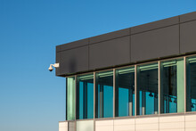 The Exterior Corner Of A Commercial Business Building. The Walls Are Dark Brown Metal Composite Panels, Tan Color Narrow Metallic Boards With Single Pane Glass Windows, And A Security Camera.