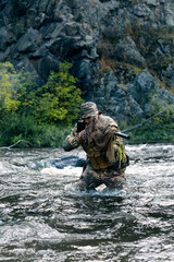 Wall Mural - Vertical photo of a mercenary military soldier during a sabotage mission behind enemy lines - he stands knee-deep in the river and aims at the enemy.