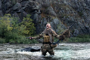 Wall Mural - Lone mercenary soldier stands knee-deep in river and screaming, arms outstretched with weapons - he lost all his friends. Despair and helplessness.