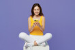 © ViDi Studio - Full body young smiling happy cool woman 20s she wear yellow tank shirt sitting on floor hold in hand use mobile cell phone isolated on plain pastel light purple background. People lifestyle concept.