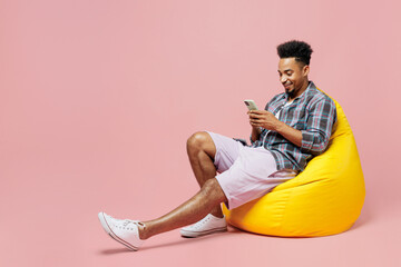 Wall Mural - Full body young man of African American ethnicity 20s he wear blue shirt sit in bag chair hold in hand use mobile cell phone isolated on plain pastel light pink background. People lifestyle concept.