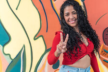 Smiling Latin American Woman Showing Peace Gesture Against Graffiti Wall