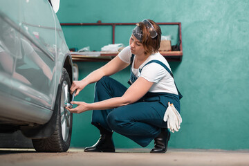 Wall Mural - Garage. Young woman mechanic in blue coveralls squatting and repair tire with ratchet wrench. The concept of women's work in men's professions