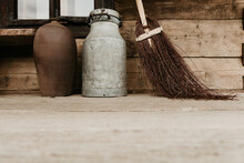 A Brown Ceramic Vase, A Vintage Metal Milk Churn Can And A Rustic Besom Broom On A Wooden Porch. Low Angle Photo With Copy Space. Countryside Home Detail Mockup. Rural Life. 