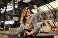 Cheerful Young Caucasian Girl Waves Her Hand Looking To Side, Spends Time At Railway Station. Blonde Woman With Phone Wears Casual Clothes In Spring. Good Mood Concept