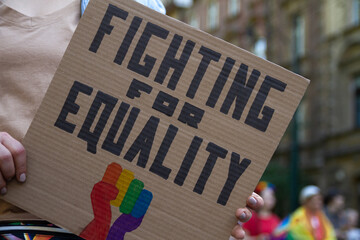 Woman holding placard sign Fighting for Equality with rainbow flag fist, during LGBT Pride Parade. Crowd of people at equality march to support and celebrate LGBT+, LGBTQ gay and lesbian community.