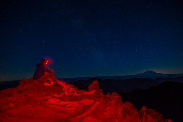 athletic adventurous male hiker sitting on top of a mountain with a red headlamp shinning, looking a
