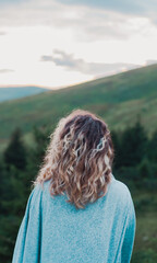 Wall Mural - girl on the background of mountains forest and sunset looks into the distance travel hike conquering the peaks from the back fair-haired curly pleasure from life prayer look into the sky to God