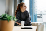 Fototapeta Panele - young redhead woman takes break in her office and looks thoughtfully to the side