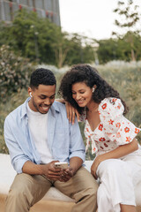 Wall Mural - Nice young african friends listen to music in headphones, look at phone sitting on street. Brunette guy wears shirt, girl wears blouse. Concept of use, leisure