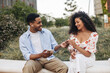 Smiling young african guy gives music to girlfriend using earphone and phone sitting on street. Couple of brunettes wear casual spring clothes. Technology concept 