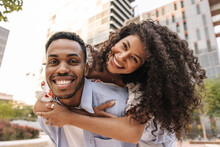Smiling African Young Girl And Her Boyfriend Are Looking At Camera Having Fun Together. Couple Of Lovers Stand In Embrace On Street. Good Mood Concept