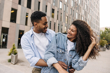 Wall Mural - Cheerful young african guy with girl look at each other relaxed walking outdoors. Couple of brunettes wear spring casual clothes. Lifestyle, different emotions, leisure concept