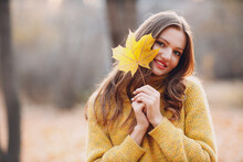 Young Woman Model In Autumn Park With Yellow Foliage Maple Leaf At Face. Fall Season Fashion