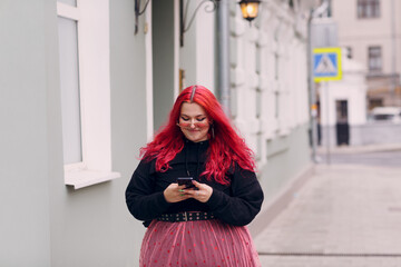 Wall Mural - European plus size woman use mobile phone outdoor. Young red pink haired body positive girl walk at city street outdoors