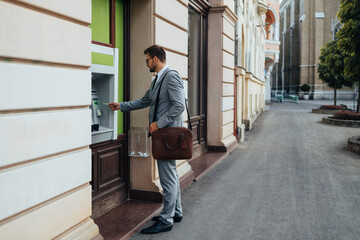 Handsome middle age businessman with eyeglasses standing on city street and using ATM machine to withdraw money from credit or debit card.