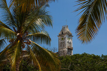 White Clock Tower Over Green Bushes And Trees And Palm Leaves In The Background Of Blue Sky. Zanzibar, Tanzania