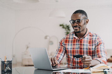 Happy african american man in glasses is purchasing online using laptop and credit card.