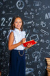 © skif - charming mischievous little girl in a school uniform, smiling at the camera, holding a magnifying glass and books in her hands, having fun during school classes against a black wall. Back to school.