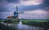 Fototapeta  - Stary wiatrak i stara zabudowa nad kanałem. Zachmurzone niebo, dramatyczne burzowe chmury. Wiejski, holenderski krajobraz.