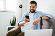 © kleberpicui - Smiling man sitting on couch at home using laptop and cell phone