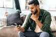 © Kateryna - Gadgets technology. Puzzled indian or arabian guy in casual wear, sits on sofa in a living room, using smartphone, texting online, browsing social networks, reads news, looks at screen in confusion