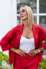 Wall Mural - Stylish fashion blonde woman in red suit and white top posing on white wall background, tropical leaves, natural daylight. Wearing trendy sunglasses.