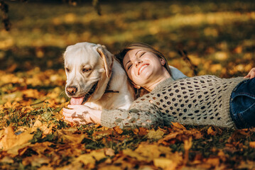 Wall Mural - A beautiful blonde lies in yellow leaves in a park and plays with her labrador.